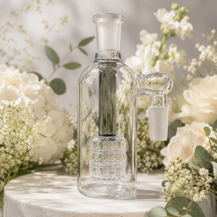 Clear glass water pipe with a frosted glass mouthpiece and inner chamber, displayed on a round stone pedestal surrounded by white flowers and greenery.