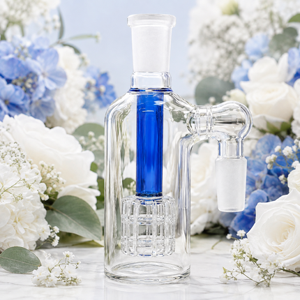 A clear glass water pipe or distiller with a bright blue inner chamber and a frosted glass mouthpiece, styled among white and blue flowers on a light marble surface.