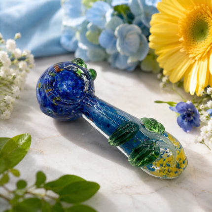 Blueberry garden glass spoon pipe displayed among blue hydrangeas, yellow flowers, and greenery on a bright marble surface