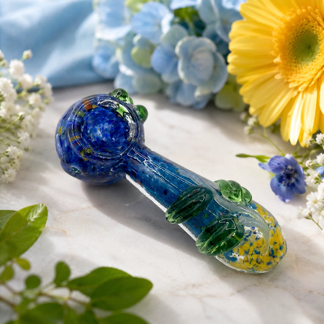 Blueberry garden glass spoon pipe displayed among blue hydrangeas, yellow flowers, and greenery on a bright marble surface