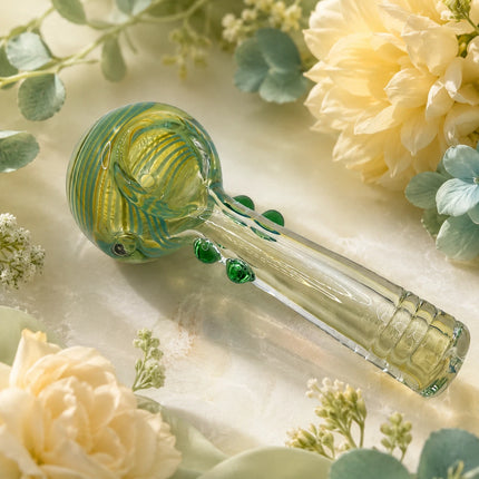 Lime and emerald glass spoon pipe arranged among soft cream flowers with green accents and a warm botanical backdrop