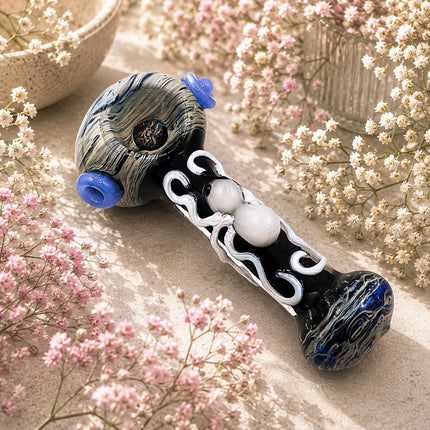 Black spider glass spoon pipe styled on a soft floral background, featuring marbled blue-gray ends and a raised white spider detail