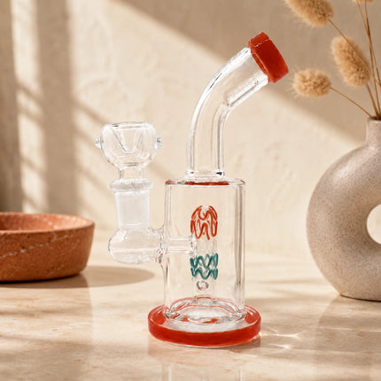 Clear mini glass bong with ruby red rim accents displayed on a warm neutral tabletop with soft natural lighting