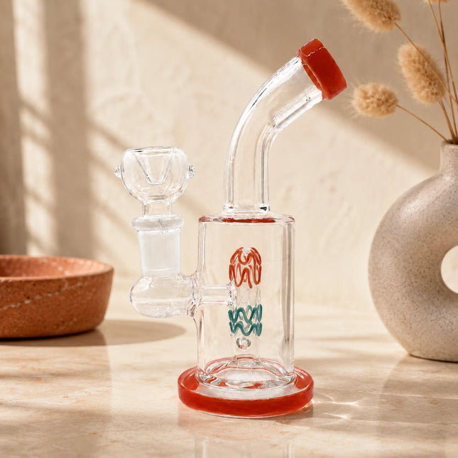 Clear mini glass bong with ruby red rim accents displayed on a warm neutral tabletop with soft natural lighting