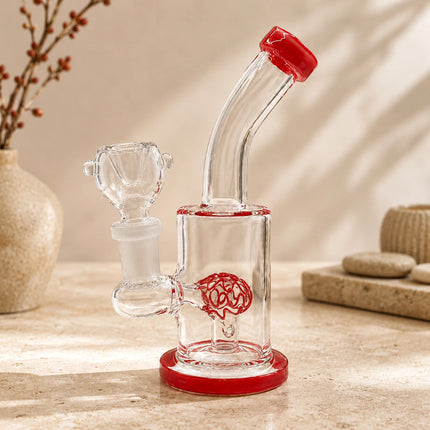 Clear red dome perc mini glass bong displayed on a neutral tabletop with soft shadows and warm natural lighting