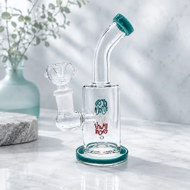 Clear mini glass bong with deep teal accents and a 14mm bowl styled on a light stone surface with a soft white floral background