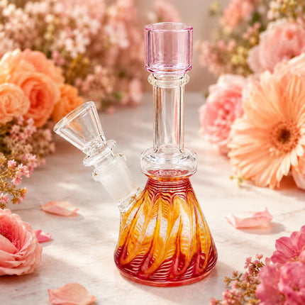 Pink sunset swirl mini beaker bong styled among peach and coral flowers on a bright decorative tabletop