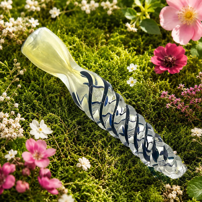 Navy spiral chillum displayed across soft green moss with pink and white garden flowers surrounding the clear glass body.