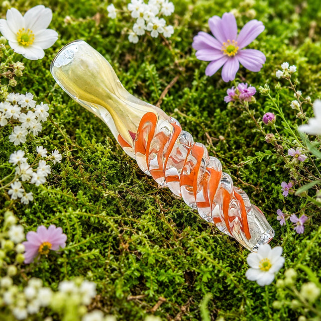 Clear 3-inch chillum styled on a mossy garden bed with bright orange spiral accents and small white and purple flowers.