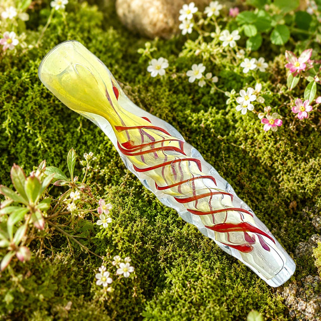 Clear chillum with red and yellow spiral accents displayed on a mossy garden surface surrounded by small white and pink flowers.