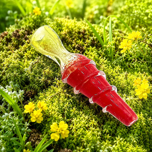 A red and translucent yellow glass smoking accessory, resembling a miniature conical flask, resting on a bed of green moss and small yellow flowers.