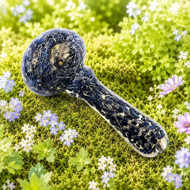 Navy blue crackle glass spoon pipe photographed outdoors among white daisies, purple flowers, and fresh green foliage.