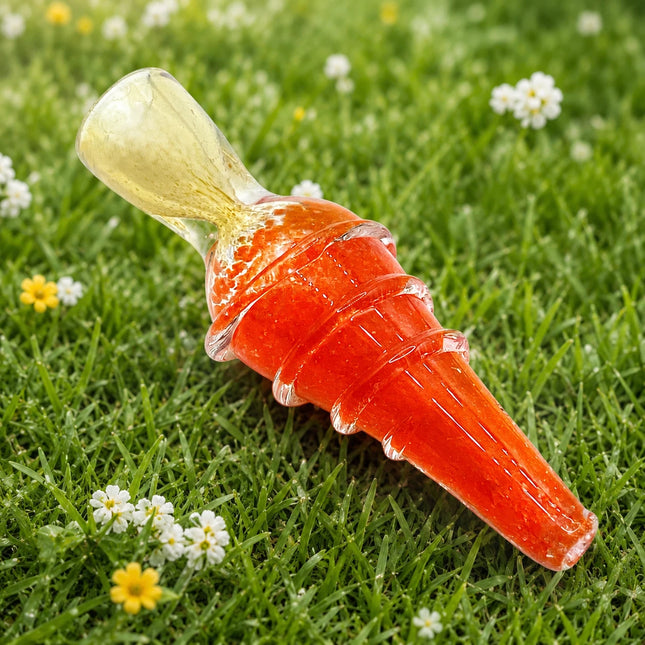 A clear glass water pipe with a vibrant orange-red body and a pale yellow-green bowl, resting diagonally on a green grassy lawn with small white and yellow flowers.
