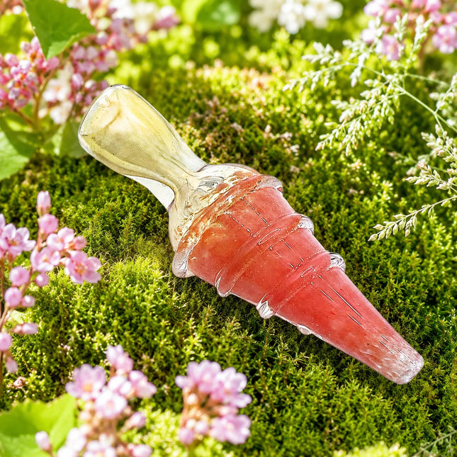 Coral pink ribbed glass chillum pipe with a pale yellow mouthpiece displayed on green moss with small pink and white flowers.