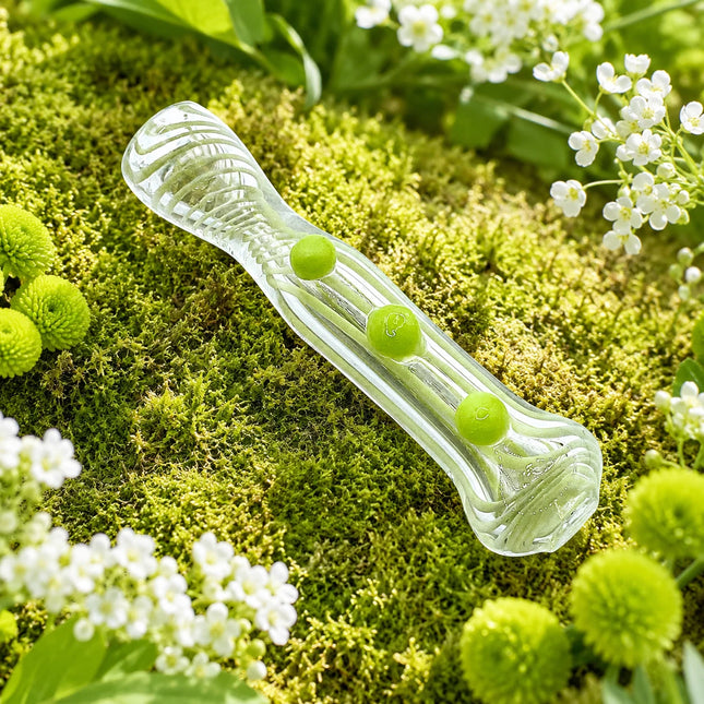 Clear 3-inch spoon pipe displayed on a fresh moss garden backdrop with bright melon green grip dots and small white flowers.
