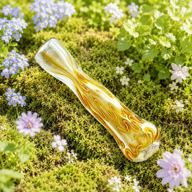 Amber swirl glass chillum pipe resting on green moss with small purple and white flowers in the background
